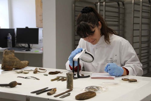 A phorensics expert studies World War One artefacts in a lab. She is using a magnifying glass and is dressed in a white lab coat and blue gloves.