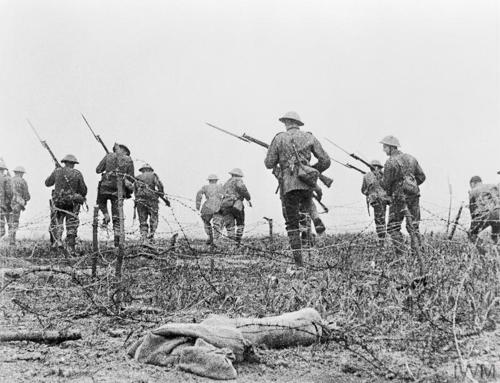 British WW1 soldiers advance into No Man's Land. The troops are wearing round Tommy helmets and carrying rifles equipped with bayonets. They are stepping over barbed wire onto muddy ground.