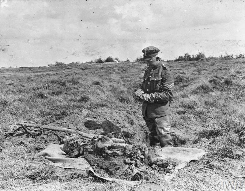 A soldier in his military uniform stands next to the exhumed body of a dead First World War infantryman. A worn rifle can be seen lying next to the remains.
