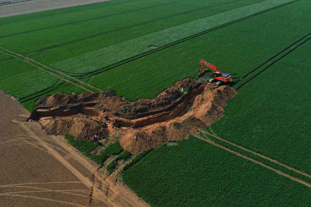 A large trench dug into a green farmer's field with earthen embankments. A red earthmover can be seen at the end of the deep trench.