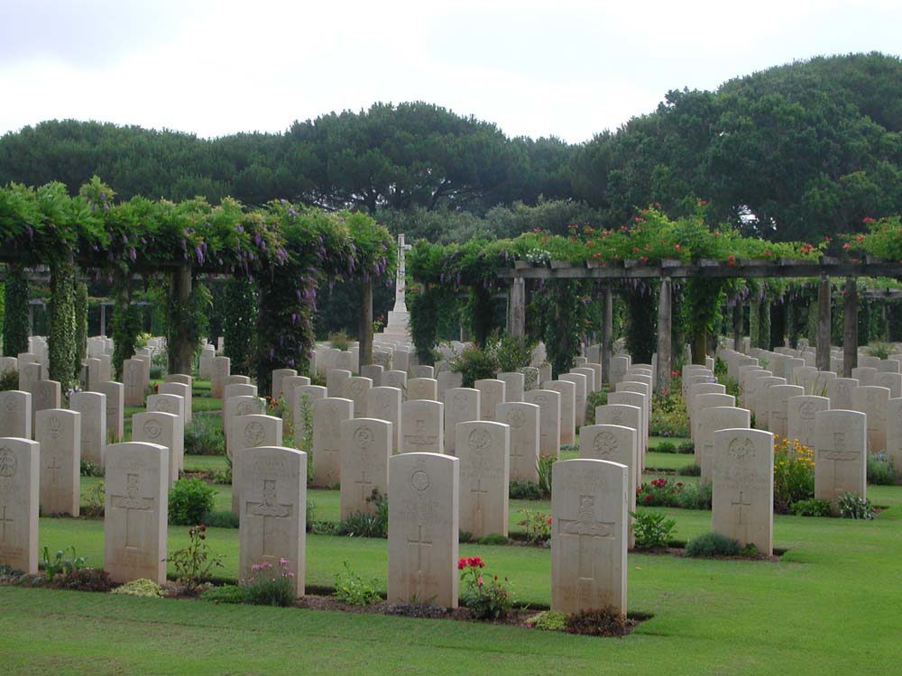 A view of Anzio Beach Head War Cemetery showing staggered rows of white rectangular CWGC headstones. Viniculture-inpsired shelters can be seen in the background, topped with foliage, while the Cross of Sacrifice is also visible in a gap between the shelters.