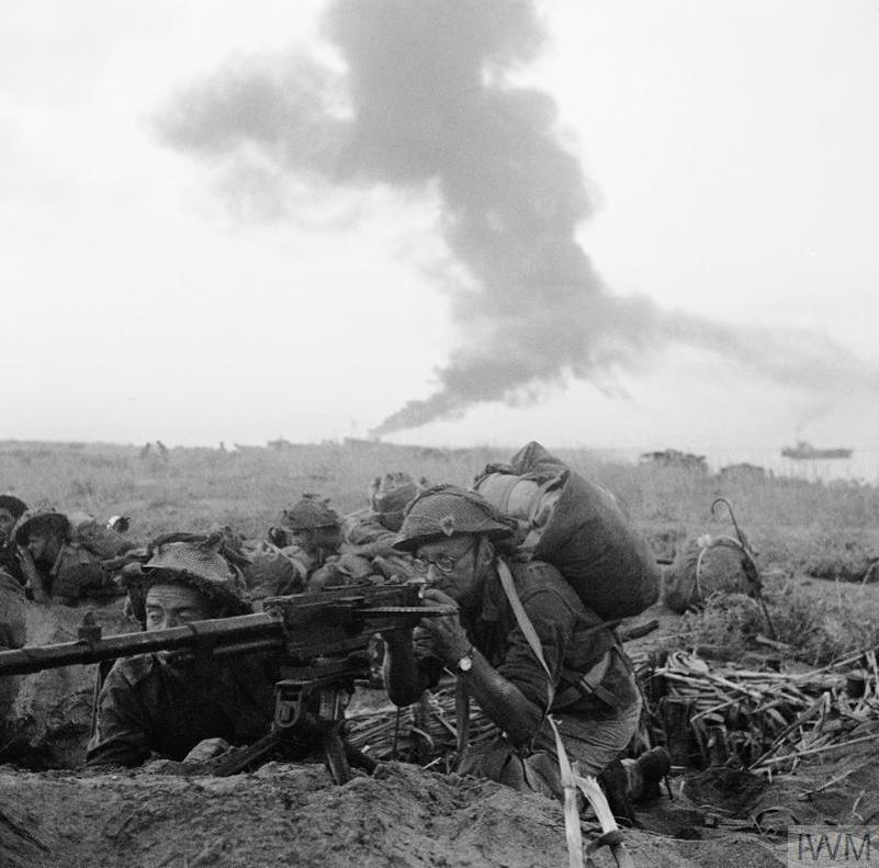 A British soldier in glasses firing a machine gun while a plomb of smoke billows into the sky behind him.