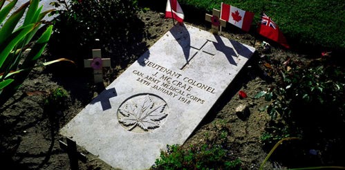 Headstone of Lt. Col John McCrae set into the ground. Red poppies on a small wooden cross adorn the graveside alongside miniature Canadian flags.
