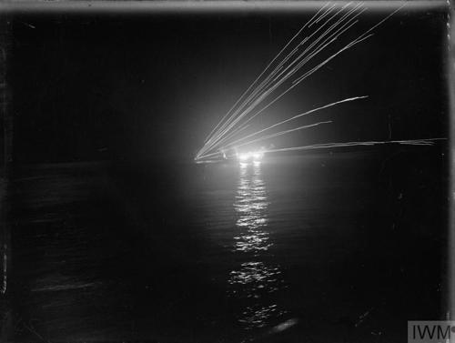 Tracer fire from shells blasting into a dark night fired out of ship's cannons. Lights dance across the water from the ship but it is too dark to see anything but the outline of the warship.
