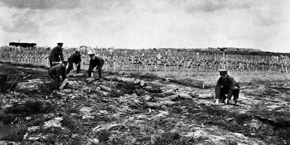 Soldiers in front row of wooden cross war grave markers while disposing of unexploded shells.