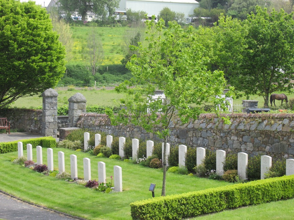 Shot showing row of CWGC headstones against a slate grey stone wall amdist the greenery of Foulon Cemetery.