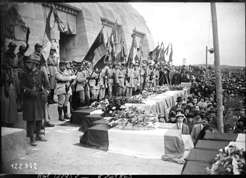 A parade of French soldiers stands in front of the oblong Douaumont Ossuary. Several porticos and entrances have been carved into the building's side. Some of the French soldiers are holding French flags in front of several coffins which have been draped in flowery wreaths and French tricolour flags.