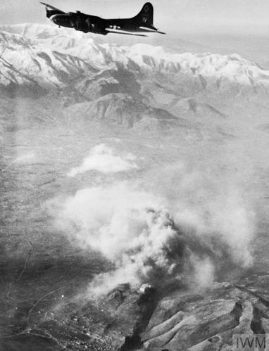 A World War Two era heavy bomber passes above Monte Cassino. A huge cloud can be seen from the explosion while in the background snowcapped mountains are visible.