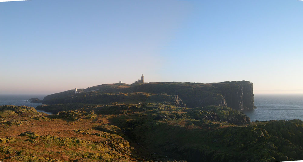 Panoramic view of the Isle of May with lighthouses in full view amidst brown/grey craggy terrain. Seabirds circle overhead.