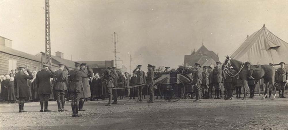 Black and white photo of the funeral procession of John McCrae. Generals salute the coffin, which is draped in a Union Jack. The coffin is sat on the back of a wheeled gun carriage. A stallion stands stoicly behind the carriage.