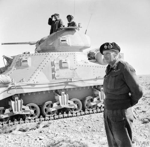 General Montgomery wearing iconic tankers beret stands next to his command tank in the desert while a crew member scans the horizon with binoculars from the tank's turret.
