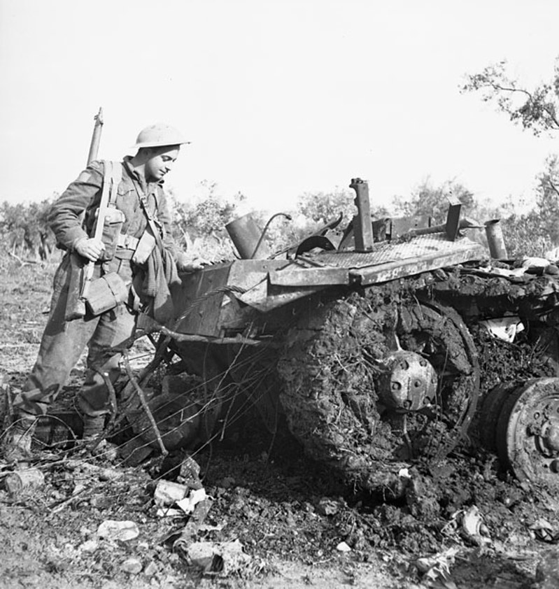 Canadian WW2 solider holding a rifle stands next to the smashed and twisted wreckage of a German vehicle.