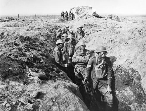 Several Australian soldiers advance through a shallow reserve trench on a WW1 battlefield. The ground has been churned up by artillery fire. The soldiers are all wearing gas masks.