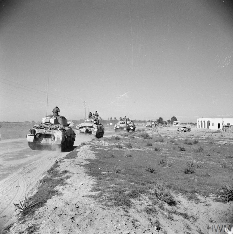 Black and white photo showing a column of Sherman tanks speeding through the desert. On the right is a small traditional dwelling.
