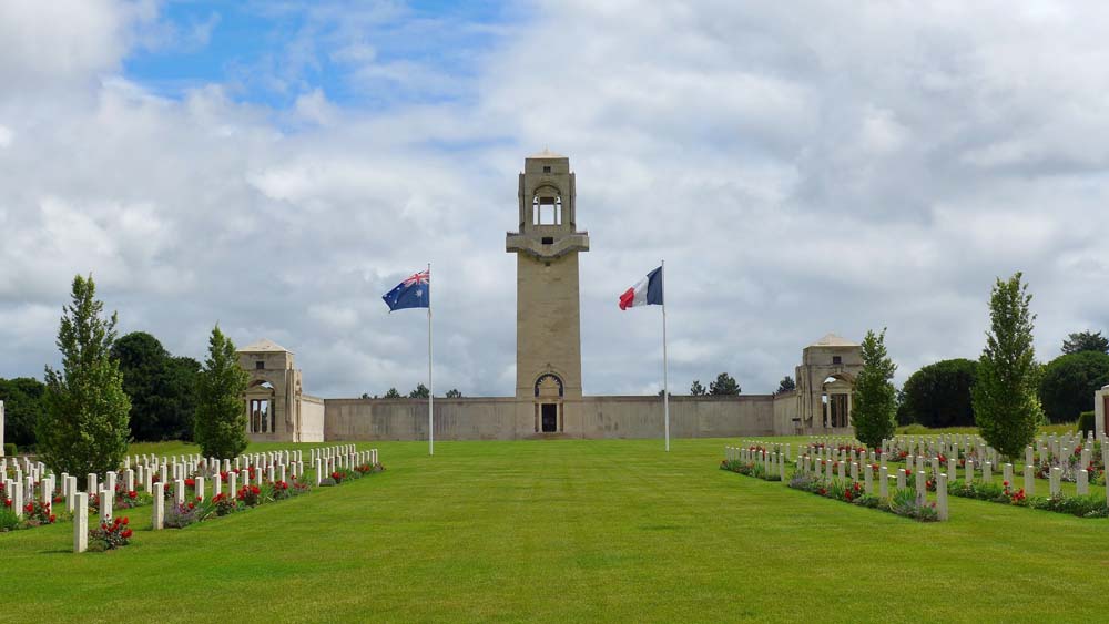 Tower of the Villers-Bretonneux Memorial sitting with rows of headstones with Australian and French flags flapping in the wind.