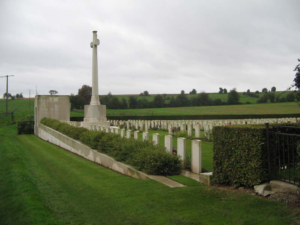 Forceville Cemetery Extension showing white headstones, Cross of Sacrifice and outhouse building offset by green lawn and fields.