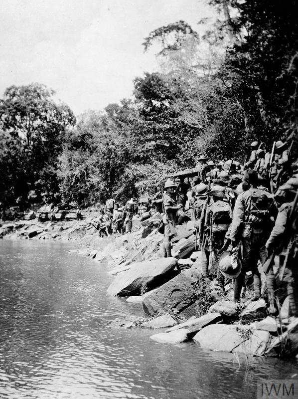 WW1 soldiers file past a river flanked by jungle.