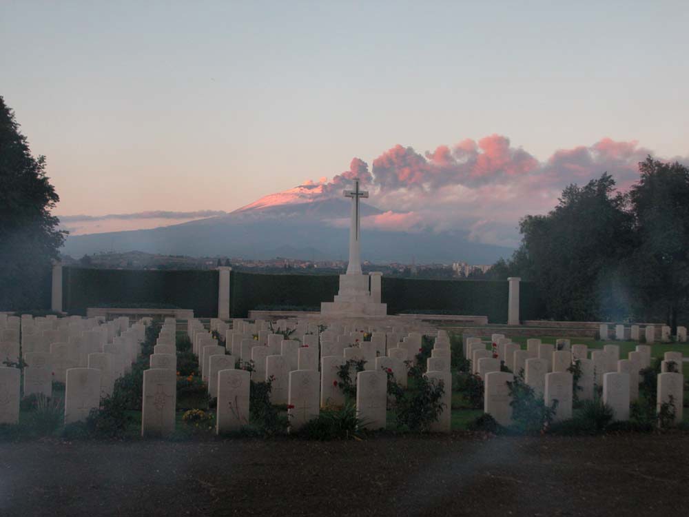 View of Catania War Cemetery showing the rows of headstones and Cross of Sacrifice in the foreground. Smoke billows from Mount Etna in the background.