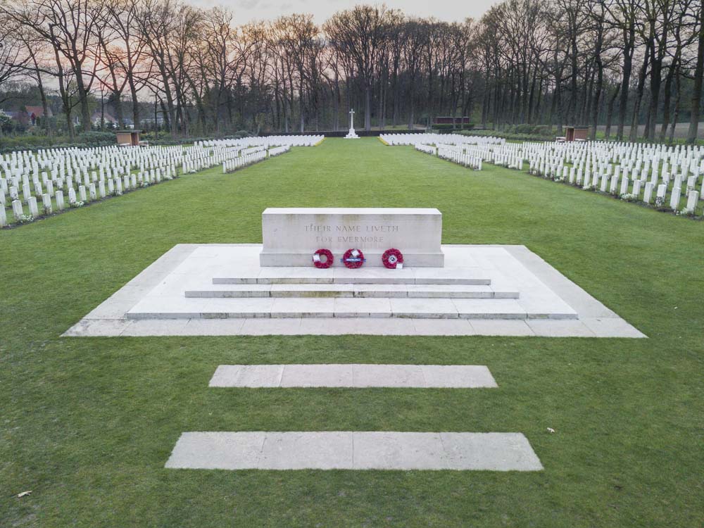 A white Stone of Remembrance on a stepped podium set amongst the greenery of Arnhem Oosterbeek War Cemetery.