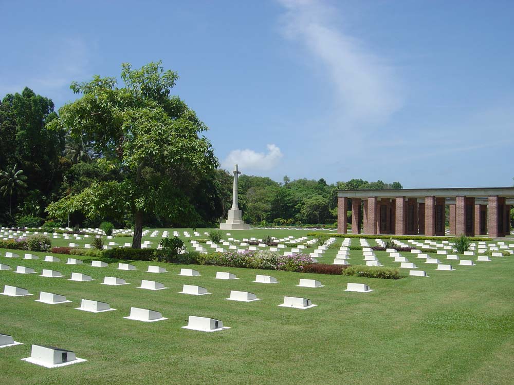 Wide shot showing the small, rectangular headstones at Labuan War Cemetery with the Cross of Sacrifice and Memorial in the background.