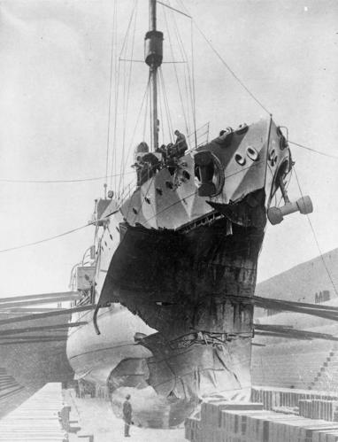 HMS Fearless in a dry dock waiting for a repairs. A huge jagged hole has been torn in the ship's bow following a collision with a submarine. An dock worker can be seen looking up at the damaged ship.