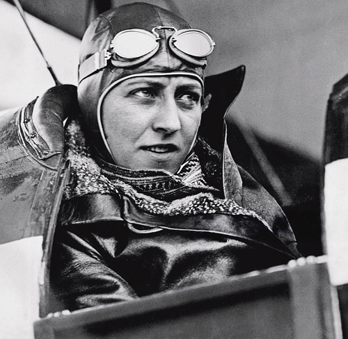 Amy Johnson in a leather flying jacket, goggles, and scarf, at the cockpit of a bi-plane.