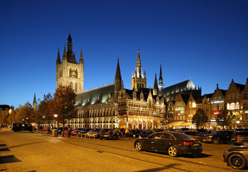 Ypres Cloth Hall on a clear evening. The sky is a vivid blue. The cloth hall displays classic medieval gothic architecture with many spires and a central clock tower.