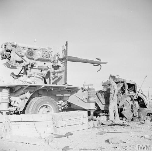 A black and white photograph showing a New Zealand soldier in trousers standing next to a knocked out Flak 88 artillery piece at the Battle of El Alamein.