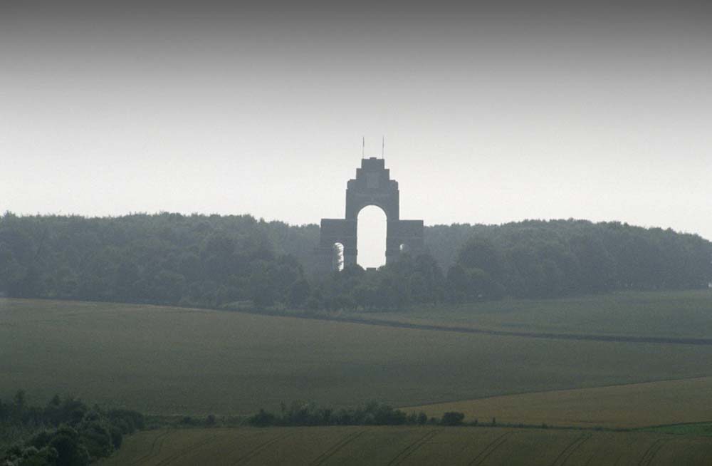Wide shot showing the triumphal arch of the Thiepval Memorial, flanked by thick woodland. Rolling farmers fields can be seen in the foreground.