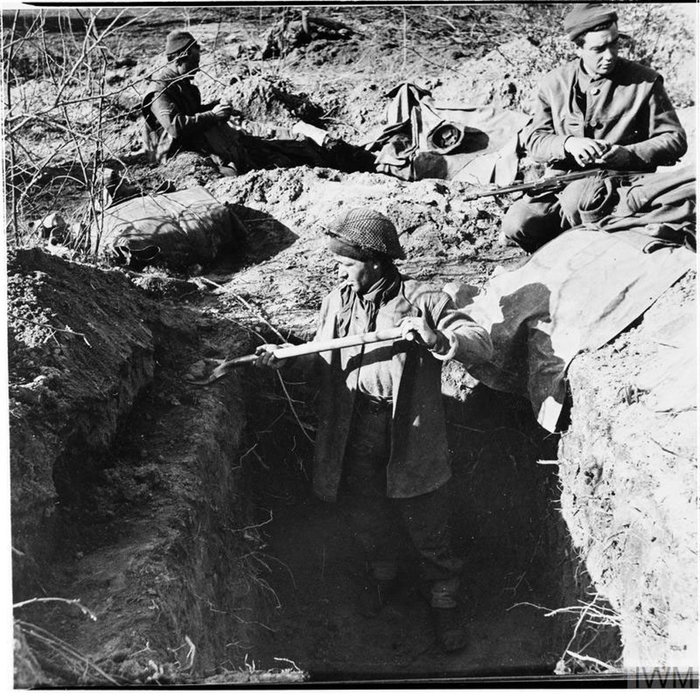 British WW2 soldiers digging a trench. Several soldiers are sitting around at the back, eating or resting, while a central soldier in the trench digs with a spade.