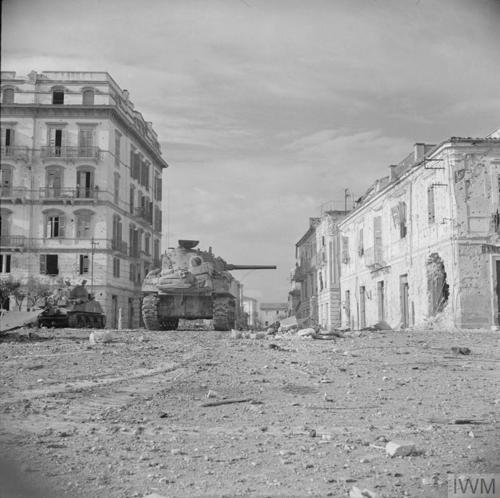 A pair of Sherman Tanks advance through the ruins of Ortona, Italy. They are passing down a wide road, flank on both sides by damaged but ornate Italian buildings.