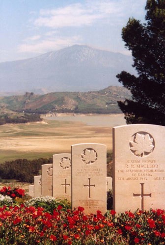 View down to Mount Etna from the top of Agira Canadian War Cemetery. CWC portland stone headstones are visible in the foreground, flanked by red flowers and blooms.