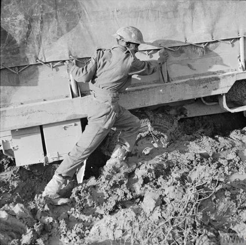 An Indian Army soldier of World War Two helps push a truck up a hill that has become stuck in heavy thick mud.