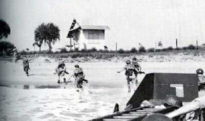 British WW2 soldiers storming up a beach in Madagascar. They have just exited a green metal landing craft. The soldiers are advancing up a beach. A wooden beach hut with a triangular roof can be seen in the background. Little shrubs and trees dot the ground.