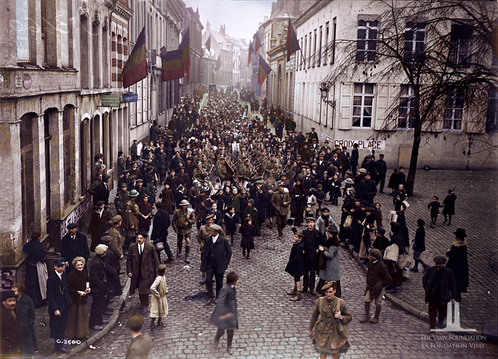 A column of Canadian soldiers marches up a street in Mons, Belgium, surrounded by civilians. Belgian flags flutter from the tall white buildings flanking the street.