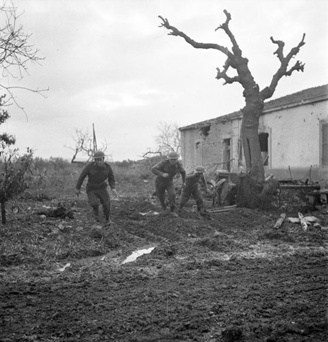 Three Canadian soldiers run for cover over a churned field next to a low farmhouse. A bare, twisted tree is visible in the foreground.